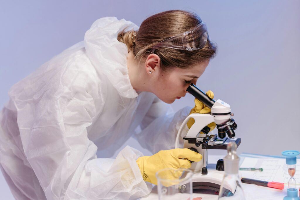 Laboratory scientist in protective gear examining samples with a microscope.